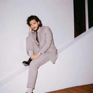 Kit Harington, actor with a full beard, dark curly hair, in a formal gray suit. White backdrop. Event promotion.