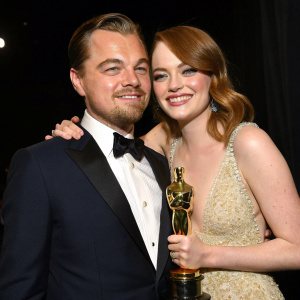 Leonardo DiCaprio and Emma Stone, posing closely together, holding Oscar statuette. Dark background, close-up shot