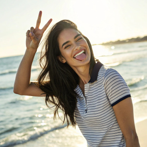 Photogenic woman with long dark hair and a cheerful smile, striking a playful pose on a sunny beach. She wears a casual striped polo shirt and gives the peace sign, sticking out her tongue