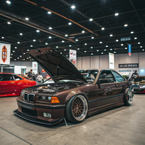 Modified BMW E36 on display at an auto show. Dark brown paint job. Open engine bay revealing high-performance engine. Indoors, well-lit venue with other cars and banners indicating theme.