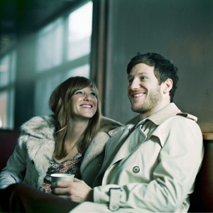 Sarah Robinson and Matt Cardle at a cozy cafe. Both smiling, Sarah with long wavy hair, wearing a fur-lined jacket and patterned top. Matt, with a closely cropped dark haircut and light stubble, dons a beige trench coat. Background blurred, warm lighting