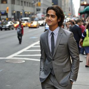 Avan Jogia, young actor, wearing a gray suit and tie. Standing in front of a busy city street. Crowds of people passing by