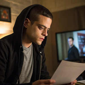Rami Malek as Elliot from Mr. Robot. Elliot, a man with short hair, wearing a black hoodie and gray shirt, intensely looking at a document in a dimly lit room. Part of another character is visible off-screen. The lighting is soft and warm, casting a subtle glow on Elliot.