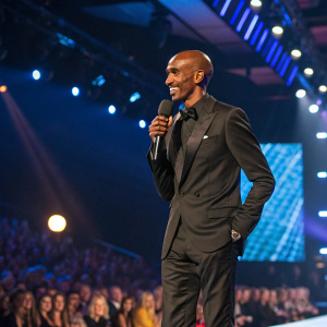 Mo Farah, British long-distance runner, in a sleek black suit, at a charity gala. Brightly lit stage, audience in background
