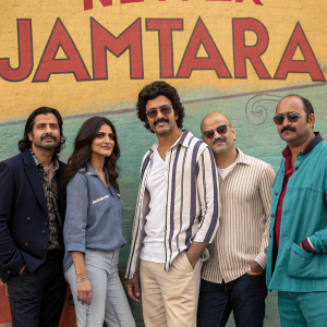 A promotional photo for Netflix series Jamtara. Five actors pose in front of a colorful backdrop with the title JAMTARA. Woman with long dark hair, balding man, man with mustache, man in striped shirt, and casually dressed man with sunglasses.