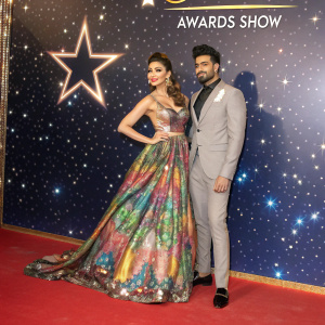 Urvashi Rautela and Gautam Gulati at a glitzy award show. The backdrop is a starlit sky with the award logo shining brightly. Both are dressed in elegant, colorful gowns and suits, respectively.