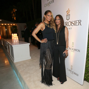 Blu Cantrell at a chic evening event, dressed in a black top and fringed pants. Luxurious Courvoisier branding in the background, alongside an unidentified woman.