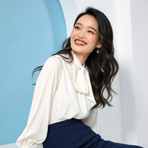 Shu Qi, with her fair skin and long wavy dark hair, wearing a sophisticated white blouse and navy skirt, smiles warmly for the camera. Simple blue backdrop