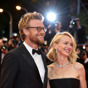 Simon Baker and Naomi Watts celebrating at Cannes Film Festival. Bright lights and bustling crowd in background.
