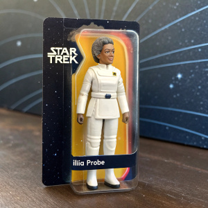 Ilia Probe toy figure in a Star Trek themed package. White uniform. Cosmic background with Star Trek logo. Collectible toy design.