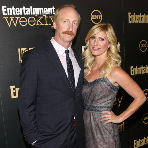 Matt Walsh and Morgan Walsh at a glitzy Hollywood event. Matt in a dark suit, mustache prominent. Morgan in a stunning dress, blonde hair flowing. Black backdrop with 'Entertainment Weekly' logos repeated.