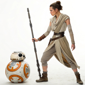 Rey, a young female hero from Star Wars, is seen in a determined pose. She has brown hair in a bun, wearing a brown and beige outfit. Standing next to a modified R2-D2 droid, on a white background.