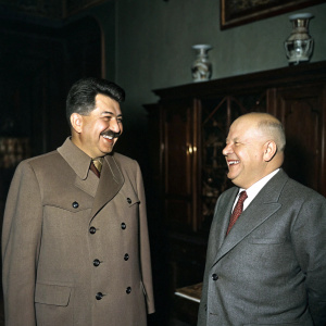 Historical photo: Joseph Stalin and Nikita Khrushchev, both in formal attire, smiling warmly. Dark, dimly lit room with old wooden furniture and vintage decorations.