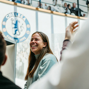 Fran Kirby, midfielder for Chelsea FC, smiling warmly. Press conference backdrop, cheering fans in the background