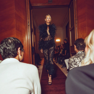 Soo Joo Park strutting down the runway at New York Fashion Week. The audience is filled with top fashion critics and industry insiders, all eyes on her intricate black ensemble adorned with a large, elegant feather.