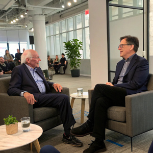 Bernie Sanders and Stephen Colbert in a casual conversation. Studio audience in the background. Modern office setting