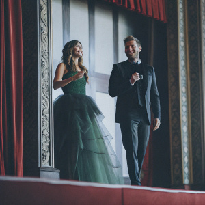 Rosie Jones and Joel Dommett attending a gala event. They stand confidently on a stage with intricate gold backdrop. Rosie in a stunning green tulle gown, Joel in a sleek black suit.