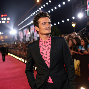 Orlando Bloom standing confidently at a high-profile awards show. He's wearing a black suit and a pink shirt with geometric patterns. The backdrop is filled with flashing lights and excited fans.