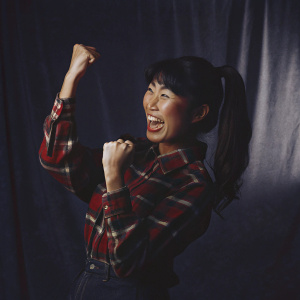 Cheerful asian woman with a ponytail, wearing a red plaid shirt, raising her fists in victory. Empty studio backdrop