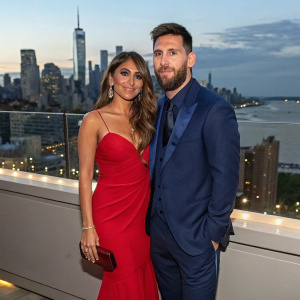Lionel Messi and Antonella Roccuzzo at the Met Gala. Both are dressed in formal attire, Messi in a navy blue suit and Antonella in a stunning red dress. The backdrop is a bustling New York City skyline.