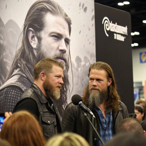 Ryan Hurst and a co-star from 'Outsiders' at a press conference. Standing in front of a large, black and white poster of the TV series. Crowded room with many attendees.
