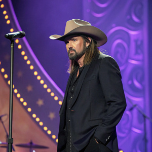 Billy Ray Cyrus in a black suit, cowboy hat, and serious demeanor. Purple backdrop with gold accents. Concert setting