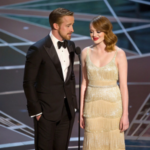 Ryan Gosling and Emma Stone at the 89th Academy Awards. They are on stage with a geometrically patterned backdrop. Ryan in a formal black tuxedo, Emma in a glamorous golden gown with layered textures.