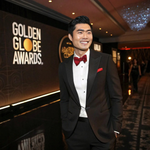 Henry Golding, Asian actor, at the Golden Globes. Styled short haircut. Wearing a classic black tuxedo with a red bow tie. Standing in a dimly lit room with a glossy floor and event logos projected on the walls.