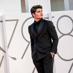 Gregg Sulkin, in a sharp black suit, stands against an abstract backdrop of geometric shapes at a gala event.