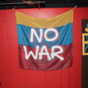 A weathered Colombian flag with graffiti 'No War' painted across it. Ripped and faded appearance, hanging on a rusty nail.