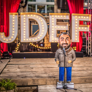 Cartoony illustration of Joe Rogan. Stands in front of a vibrant red curtain with 'JDEF' letters shining brightly on the stage backdrop