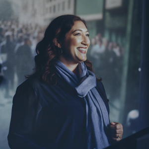 Randi Zuckerberg, beaming with a warm smile, in a sleek black blouse paired with a blue scarf. SiriuxXM promotional event with a bustling crowd in the background.