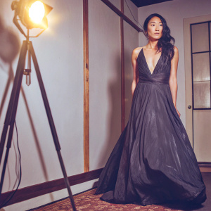Greta Lee, an Asian actress, stands in a stark white studio. She wears a flowing, black gown with a deep V-neck. Her expression is contemplative. The background is empty, with a single spotlight casting a golden glow