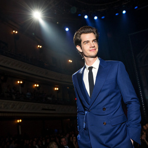 Actor Andrew Garfield at the Gotham Awards. Wearing a deep blue suit with a white shirt and black tie. Poses confidently in front of a dimly lit stage with spotlights and audience in the background.