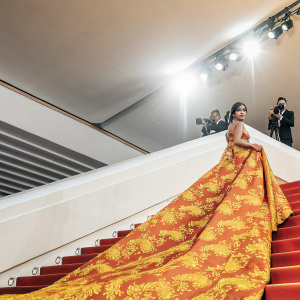 Priyanka Chopra at Cannes Film Festival. Dressed in a red and gold embroidered gown, posing confidently. Lavish red carpet scene with paparazzi and flashing lights.