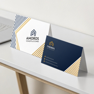 Two business cards for Amorós Construcciones, featuring a modern logo and minimalist design. One card has traditional layout with text and contact info, the other has a striped pattern. On a simple white desk.
