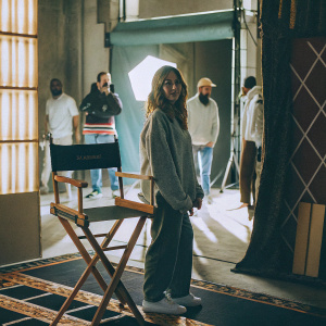 Sarah Michelle Gellar on the set of The Wonderful Maladys. She stands beside a director's chair with a unique label. The backdrop is a bustling film studio with crew members in the background.