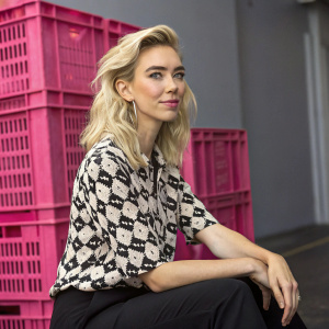 Vanessa Kirby, blonde actress, sitting confidently with a slight smile. Wearing a black and white patterned blouse. Behind her, a large pink crate.