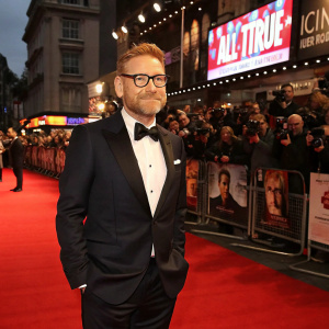 Kenneth Branagh, wearing a black tuxedo and glasses, standing on a bustling red carpet. Crowds of fans and photographers. The event is promoting the film 'All Is True'.