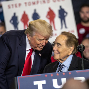 Photograph at political rally featuring Donald Trump and Bob Dole. Trump, an adult male with blonde hair, leans slightly towards Bob Dole, an older male with a friendly expression. Behind them, a banner displays silhouetted figures of people.