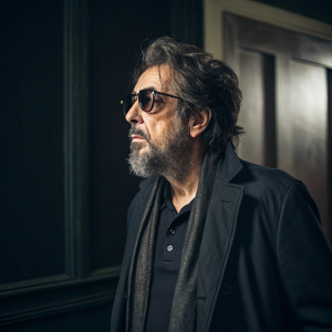Al Pacino, sporting a rugged beard, dark glasses. Standing alone in dimly lit room, against dark background.