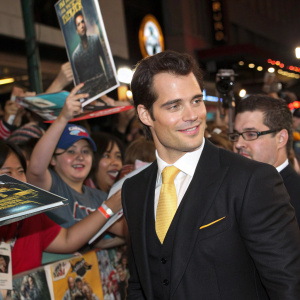 Henry Cavill at a bustling comic-con convention. Wearing black suit, crisp white shirt, and vibrant yellow tie. Surrounded by fans in casual and formal attire.