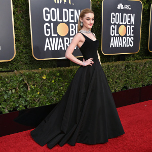 Kathryn Newton at the Golden Globe Awards. Wearing a sleek black dress with a fitted bodice and floor-length skirt. Elegant pose. The backdrop features gold accents and red carpet leading up to the stage.