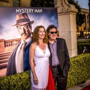 Eila Mary Matt and Barry Williams attending the star-studded premiere of 'Mystery Man' at Hollywood's El Capitan Theatre. Eila in a stunning white dress with a red skirt, Barry in a classic dark suit. The backdrop features a large poster of the enigmatic protagonist in sunglasses and hat.