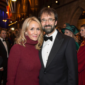 J.K. Rowling and Neil Murray at a wizard-themed charity gala. Rowling in a cozy burgundy sweater, Murray in a classic black tuxedo. Crowd in the background.