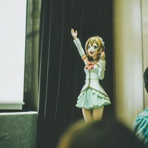 Close-up of Honoka Kousaka, animated character. Cheerful, youthful face with bright expression. Posed in dynamic stance against black backdrop