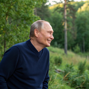 Vladimir Putin in a casual setting. Wearing a navy blue sweater. Smiling outdoors with greenery in the background.