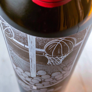 Close-up of Stoudemire Private Collection wine bottle. Black and white illustration of basketballs, NBA logos, red tinted cap. High resolution photo, focus on textures and details.