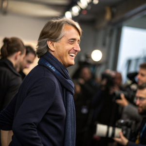 Roberto Mancini, cheerful expression, dressed in a navy sweater and scarf. Crowded backstage area with blurred photographers