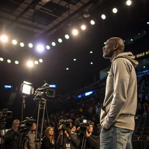 Kevin Garnett, in a casual hoodie and jeans, at a press conference. Bright lights, multiple cameras, and a packed auditorium